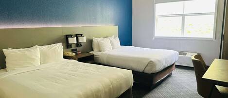 A hotel room with two beds, a desk, and a chair. There is a window with a view of greenery outside.