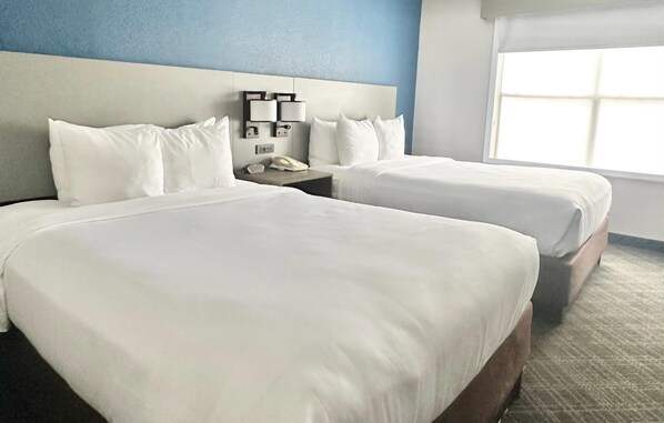 A hotel room with two beds, a headboard with wall-mounted lamps, a telephone, and a window with blinds.