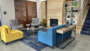 Een moderne woonkamer met een blauwe bank, een gele fauteuil en een grijze fauteuil. Er is een open haard, een boekenkast en een trap.
