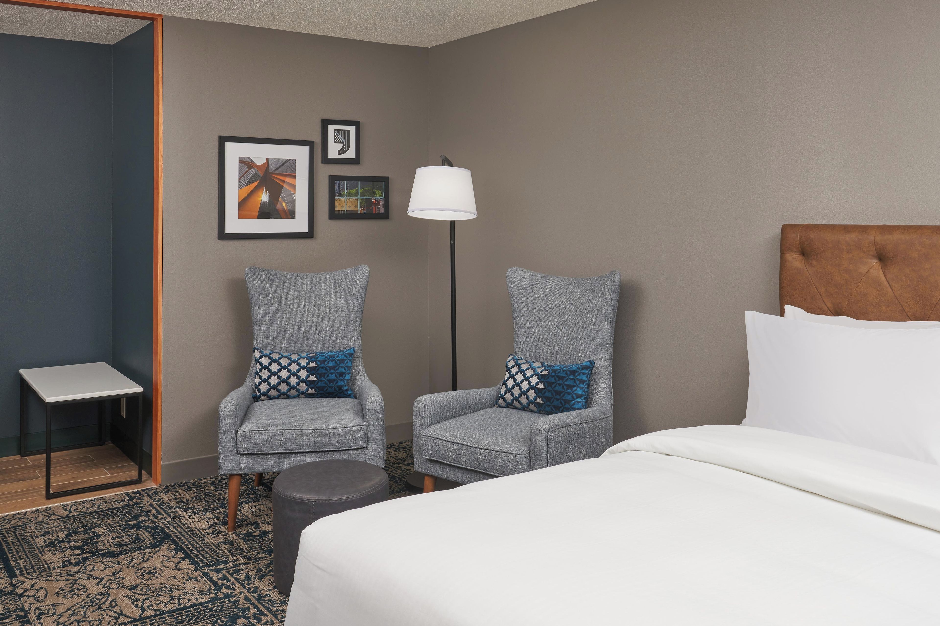 Habitación de hotel con dos sillones grises, una mesita pequeña, una lámpara de pie y una cama con ropa blanca.