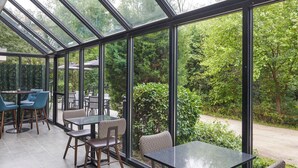 Un área de patio techada con mesas y sillas, amplios ventanales de vidrio y vistas a zonas verdes.