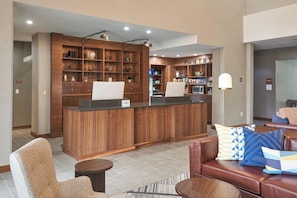 Un hall de hotel con una recepción, una zona de estar con sofá y sillones, y una kitchenette.