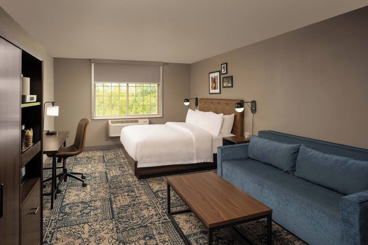 A hotel room with a bed, a blue sofa, a wooden coffee table, a desk with a chair, and a window with a view of trees.