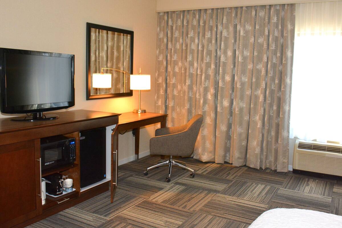 A hotel room with a flat-screen TV, a desk with a lamp, a chair, a microwave, and a bed.