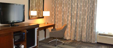 A hotel room with a flat-screen TV, a desk with a lamp, a chair, a microwave, and a bed.