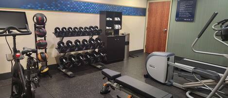 Academia com equipamentos de ginástica, incluindo esteira, bicicleta ergométrica, supino e um suporte com halteres.