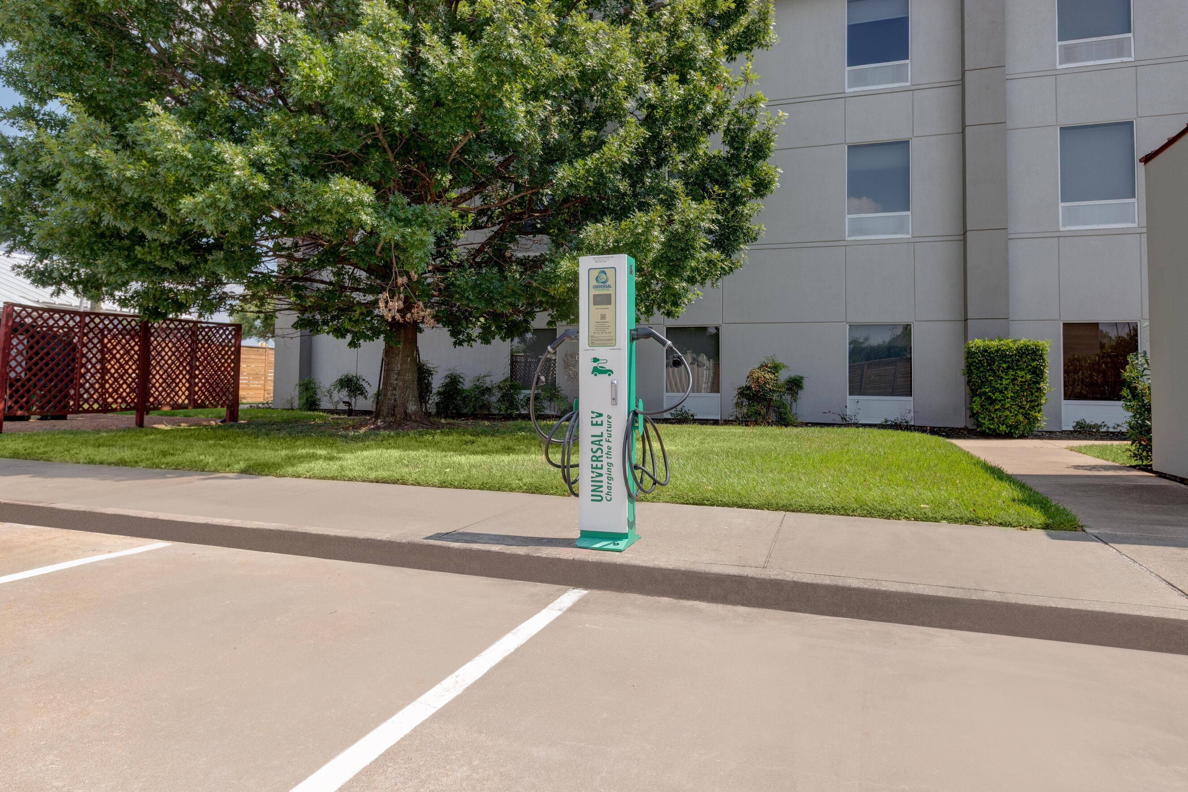 A parking area with a green electric vehicle charging station in front of a building with multiple windows and a tree.