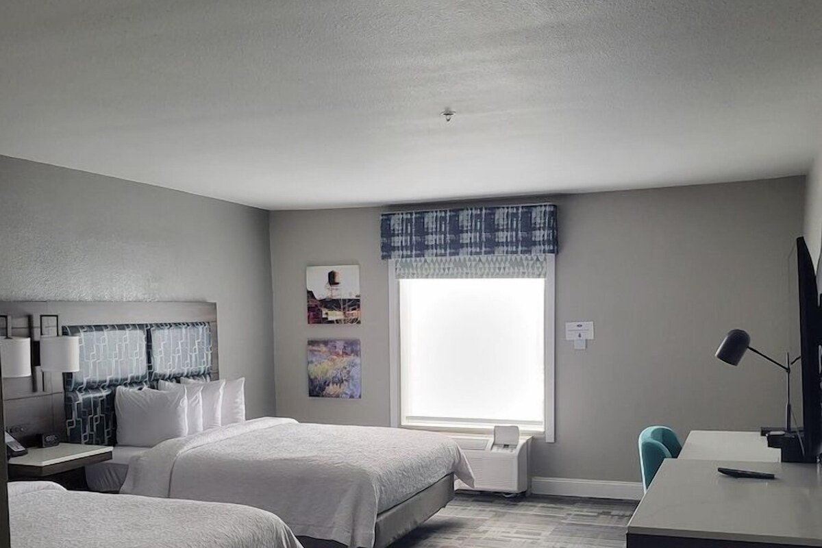 A hotel room with two beds, a desk, a chair, a window with a view, and a painting on the wall.