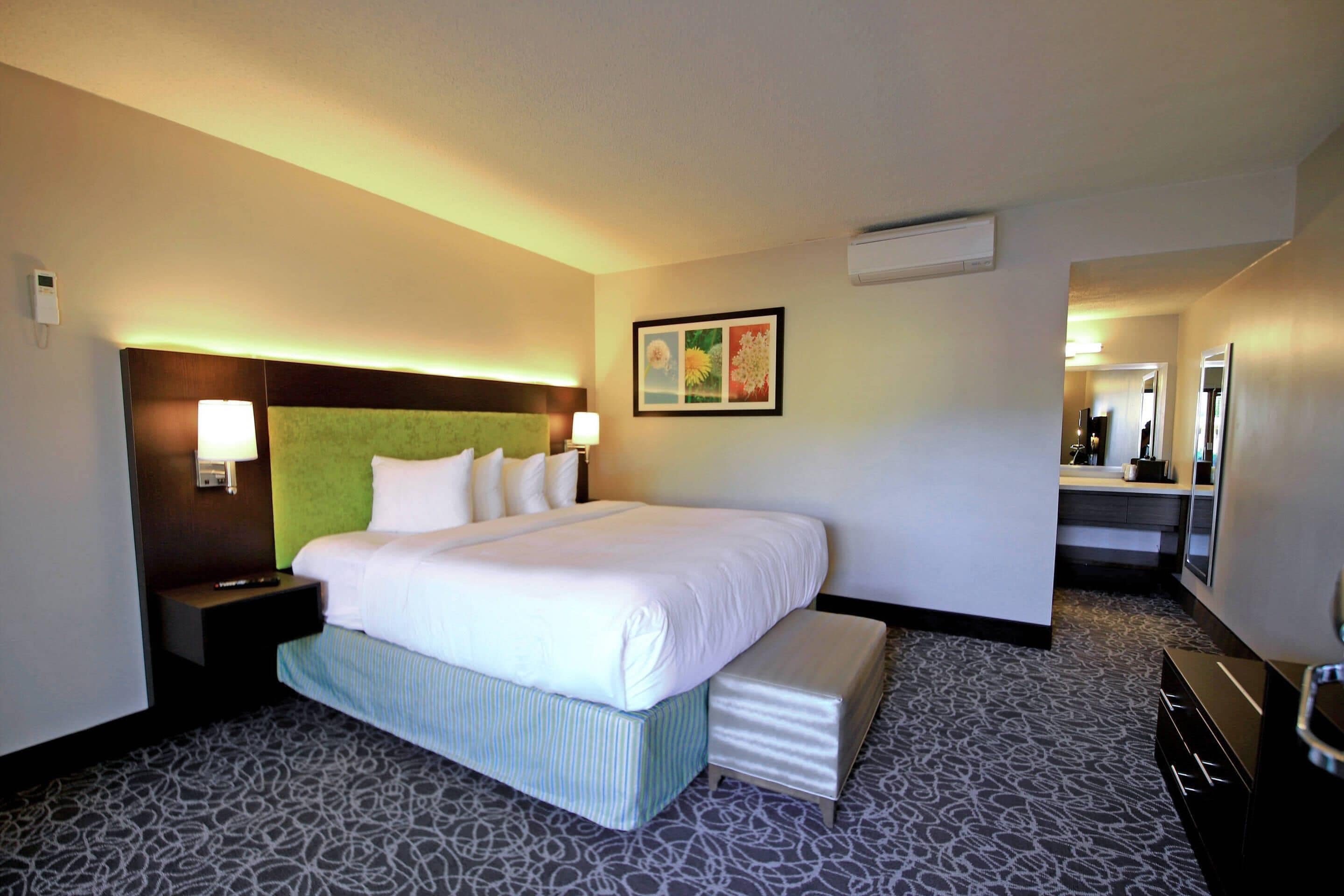 Une chambre d’hôtel avec un grand lit, des tables de chevet, une tête de lit, un climatiseur fixé au mur et un petit bureau avec une lampe.