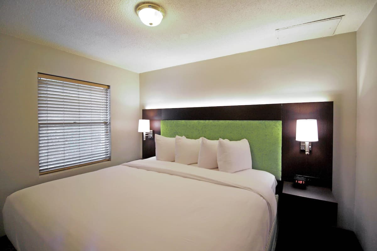 A hotel room with a large bed, a green upholstered headboard, two bedside lamps, and a digital clock on a nightstand.