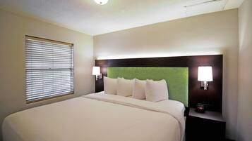Habitación de hotel con una cama grande, cabecera tapizada de verde, dos lámparas de noche y un reloj digital sobre una mesita de noche.