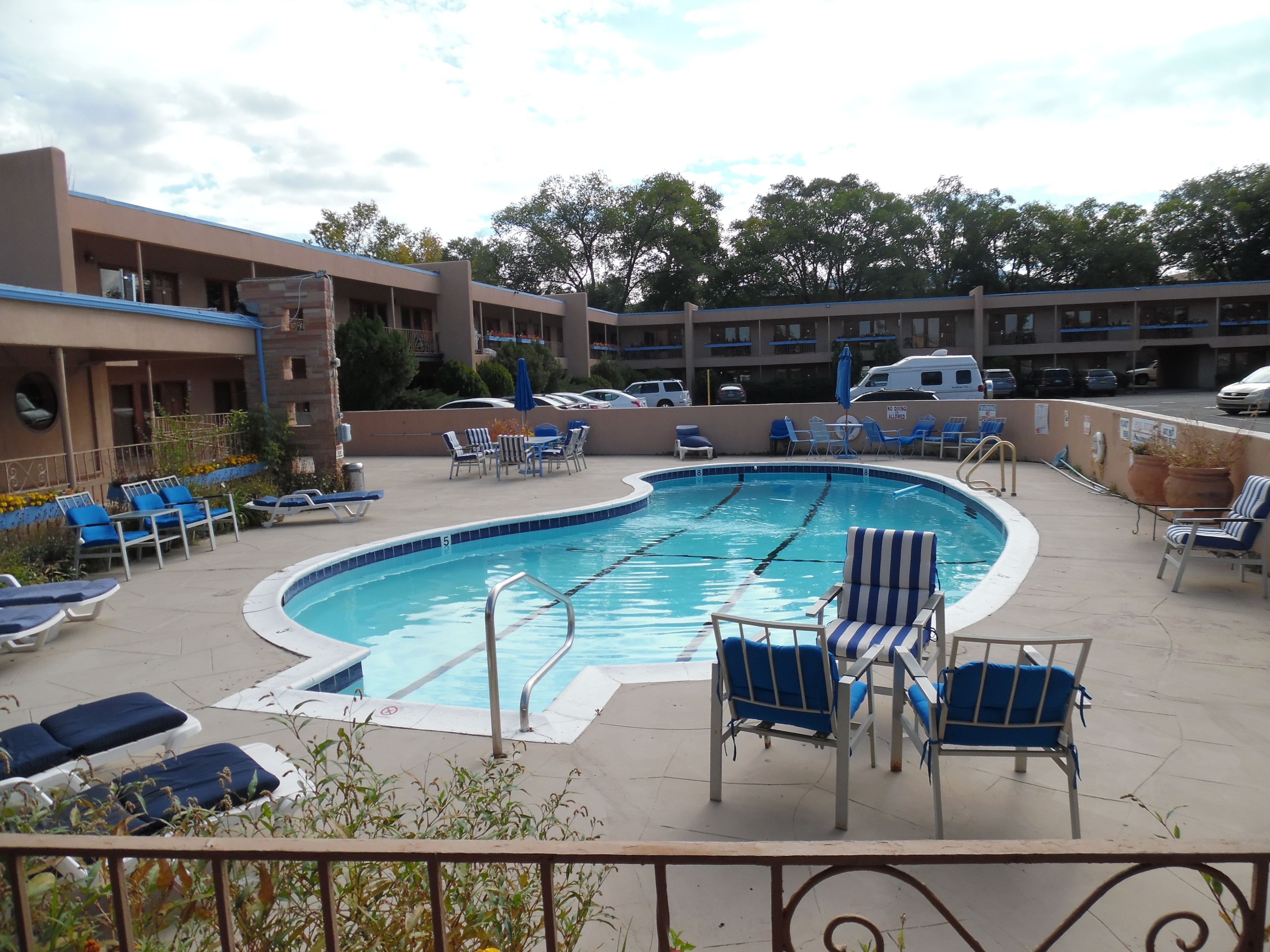A swimming pool with lounge chairs and umbrellas, surrounded by a building with multiple balconies and parked cars.