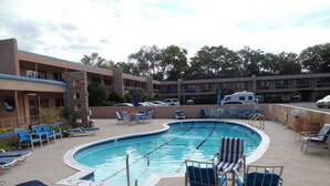 A swimming pool with lounge chairs and umbrellas, surrounded by a building with multiple balconies and parked cars.