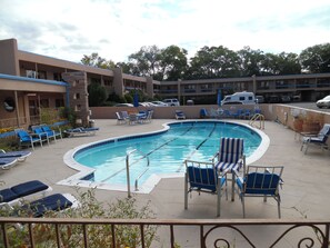 A swimming pool with lounge chairs and umbrellas, surrounded by a building with multiple balconies and parked cars.