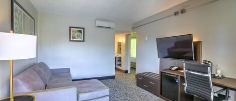 A hotel room with a sofa, a desk with a TV, a lamp, and a patterned carpet.