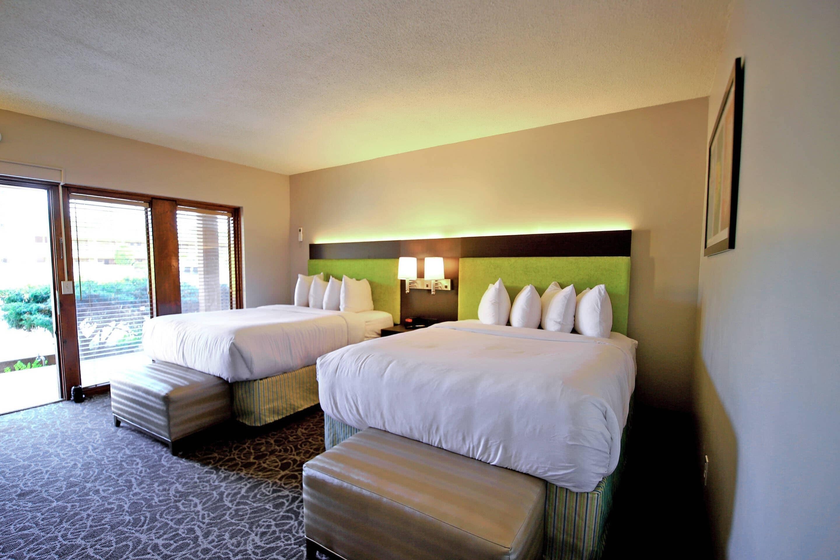 A hotel room with two beds, a bench, a door to a balcony, and a wall-mounted lamp.