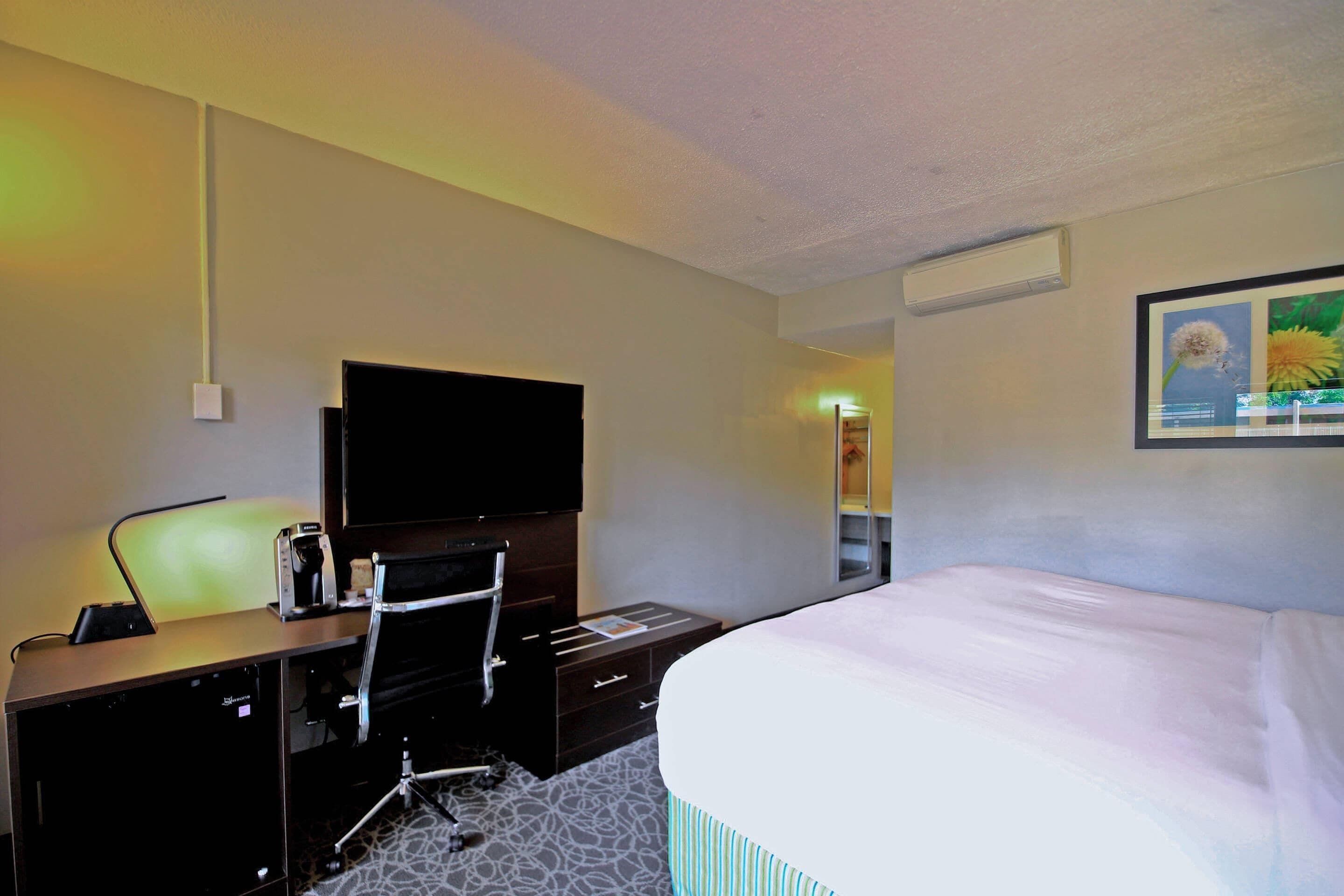 Une chambre d’hôtel comprenant un lit, un bureau avec un téléviseur à écran plat, une chaise, une machine à café et un tableau encadré au mur.