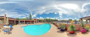 A swimming pool with lounge chairs and umbrellas, surrounded by a building with multiple windows and a covered patio area.