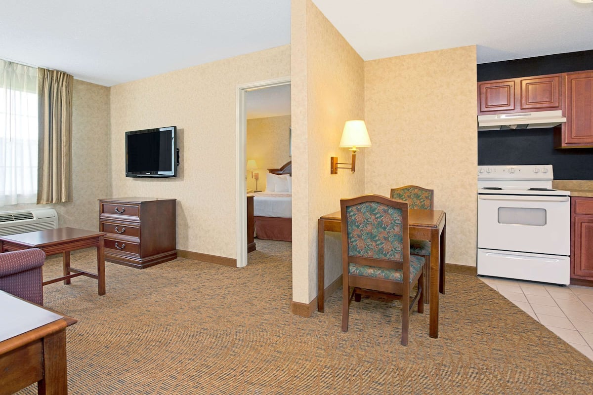 A hotel room with a bed, a desk, a chair, a television, a lamp, a dresser, and a kitchen area with a stove.