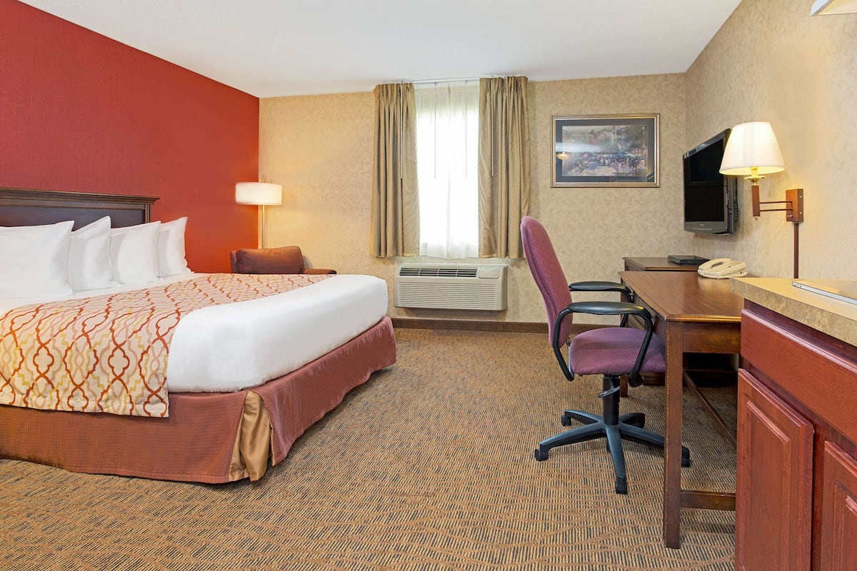 A hotel room with a large bed, a desk, a chair, an air conditioning unit, a television, and a painting on the wall.