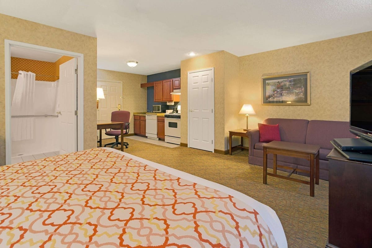 A hotel room with a bed, a sofa, a desk, a TV, a kitchen area, and a bathroom.
