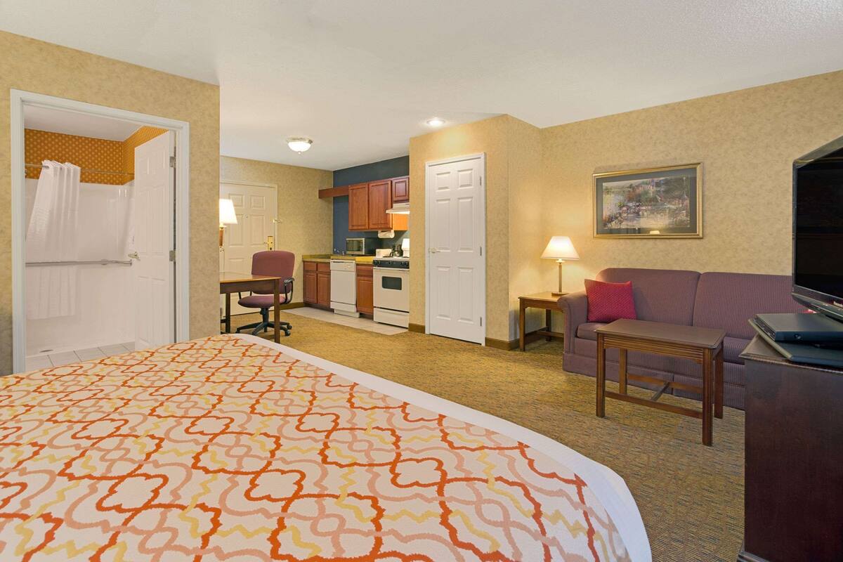 A hotel room with a large bed, a sofa, a TV, a desk, a chair, a small table, a lamp, a microwave, a stove, and a door leading to a bathroom.
