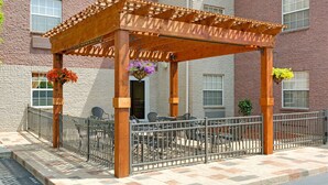 Una pérgola de madera con barandilla metálica, mobiliario exterior y macetas colgantes de flores.