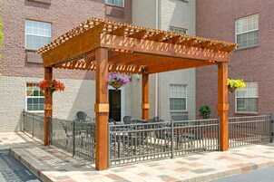 Una pérgola de madera con barandilla metálica, mobiliario exterior y macetas colgantes de flores.
