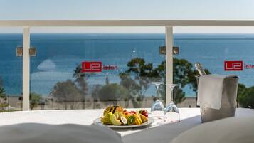 A table with a fruit platter, a glass of water, and a glass of wine, with a view of the ocean and trees in the background.