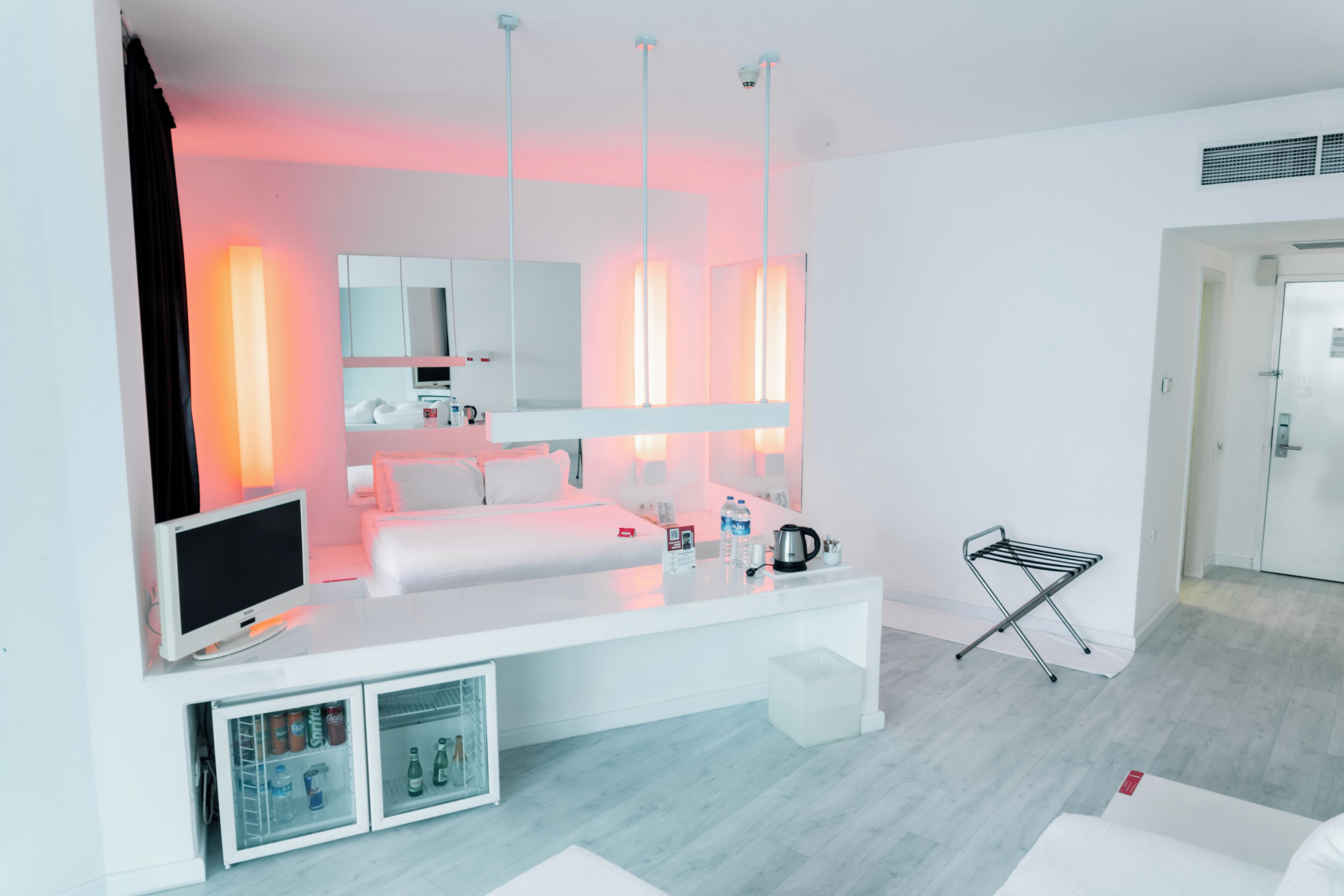 Ein modernes Hotelzimmer mit einem weißen Bett, einem Fernseher, einer Minibar und einem großen Spiegel.
