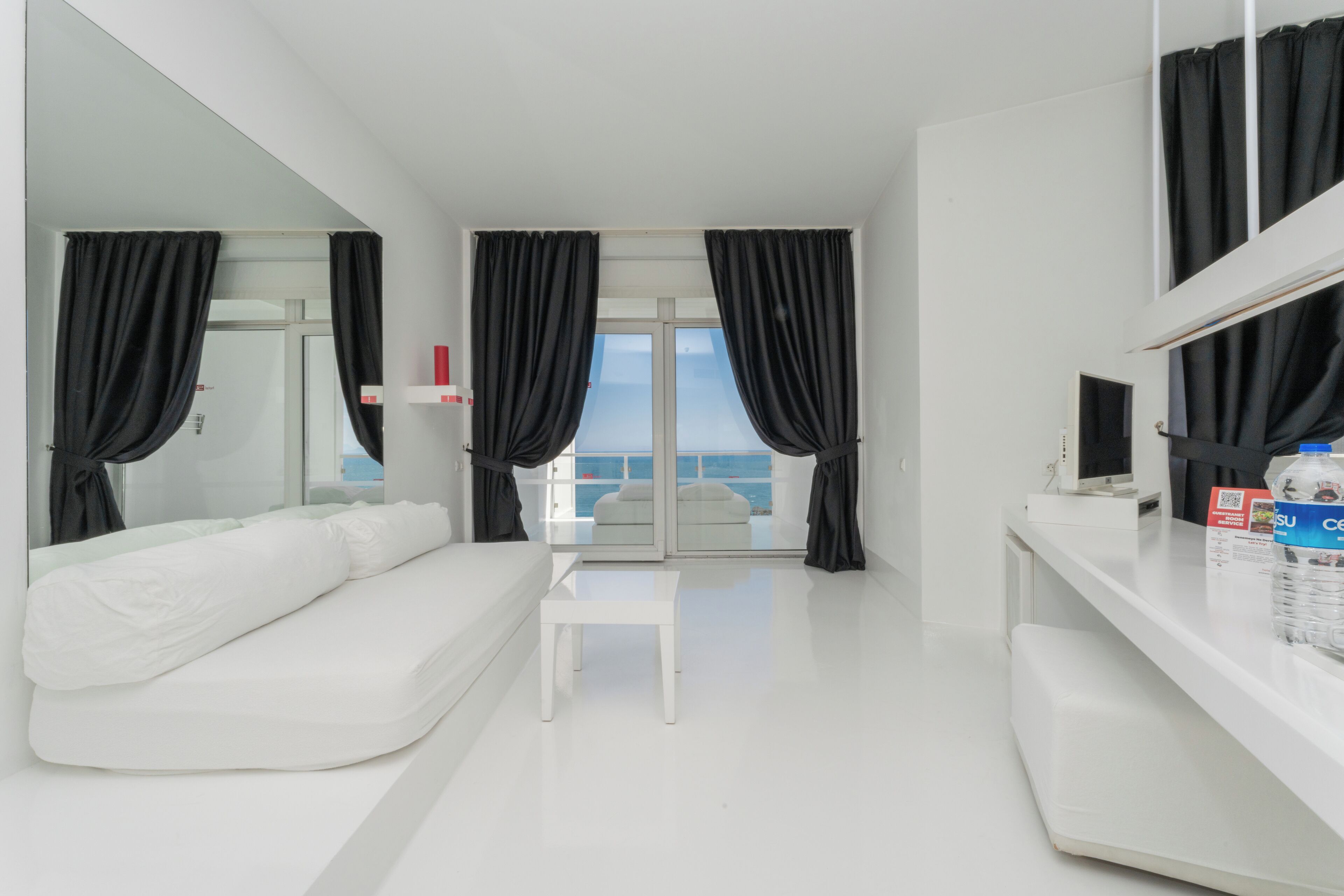 A modern hotel room with a white sofa, a small white table, and a view of the sea through large windows with black curtains.