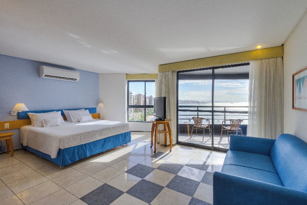 A hotel room with a bed, a TV, a balcony with a view, and a blue sofa.