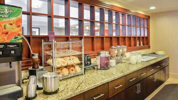 A hotel breakfast area with a coffee machine, a fruit juice dispenser, and a variety of pastries and baked goods displayed in a glass case.