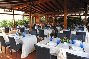 An outdoor dining area with tables set for four, covered by a wooden pergola, surrounded by chairs and plants.