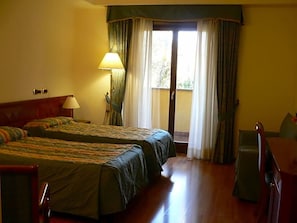 A hotel room with two beds, a desk, a lamp, and a view of the outdoors.