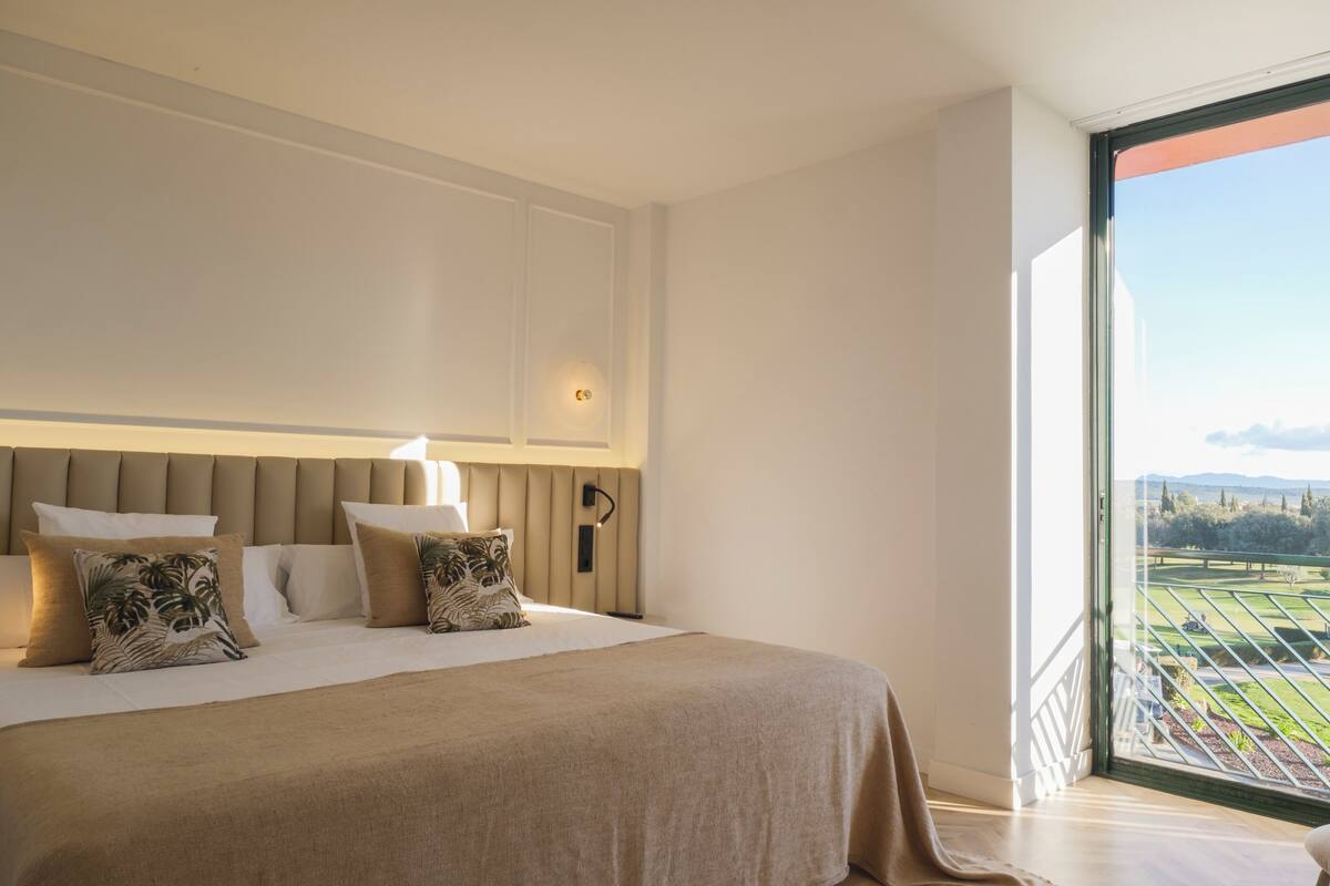 A bedroom with a large bed, a headboard with integrated lighting, a nightstand, and a view of a green landscape through a large window.