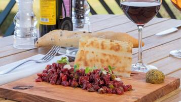 Un tavolo di legno con un pasto, inclusa una coppa di vino rosso, una bottiglia d'olio d'oliva e una bottiglia di vino, con uno sfondo panoramico.