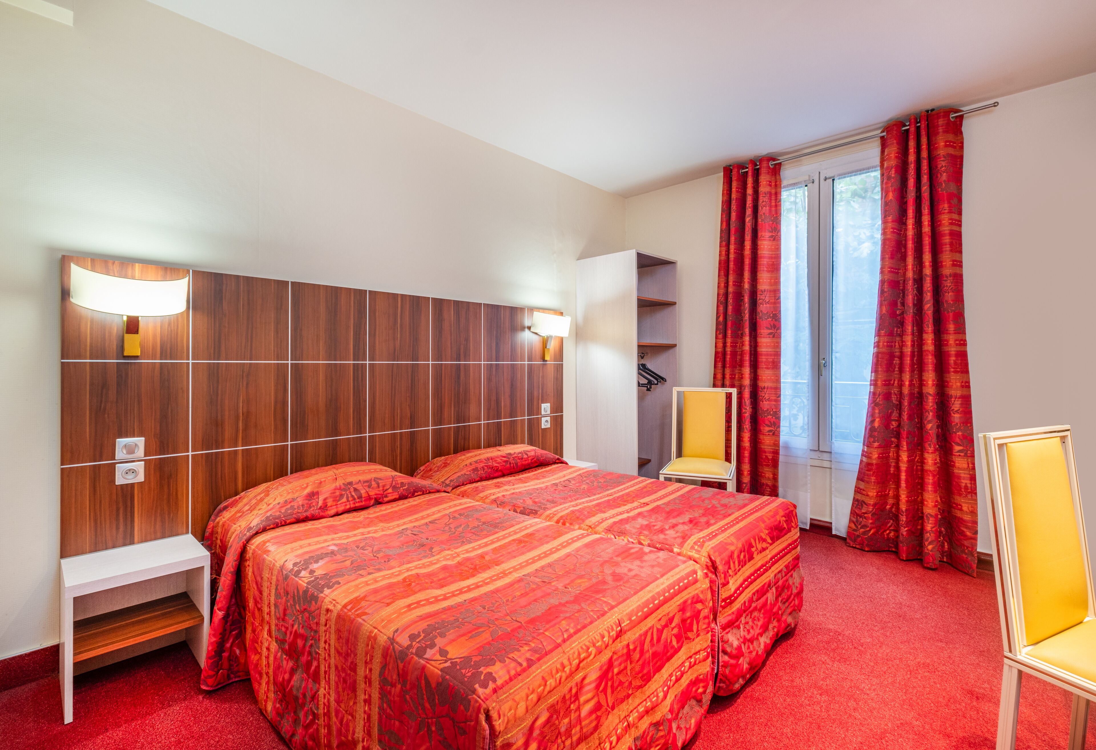Une chambre d’hôtel avec deux lits, une tête de lit en bois, une petite table, une chaise et une fenêtre avec des rideaux rouges.