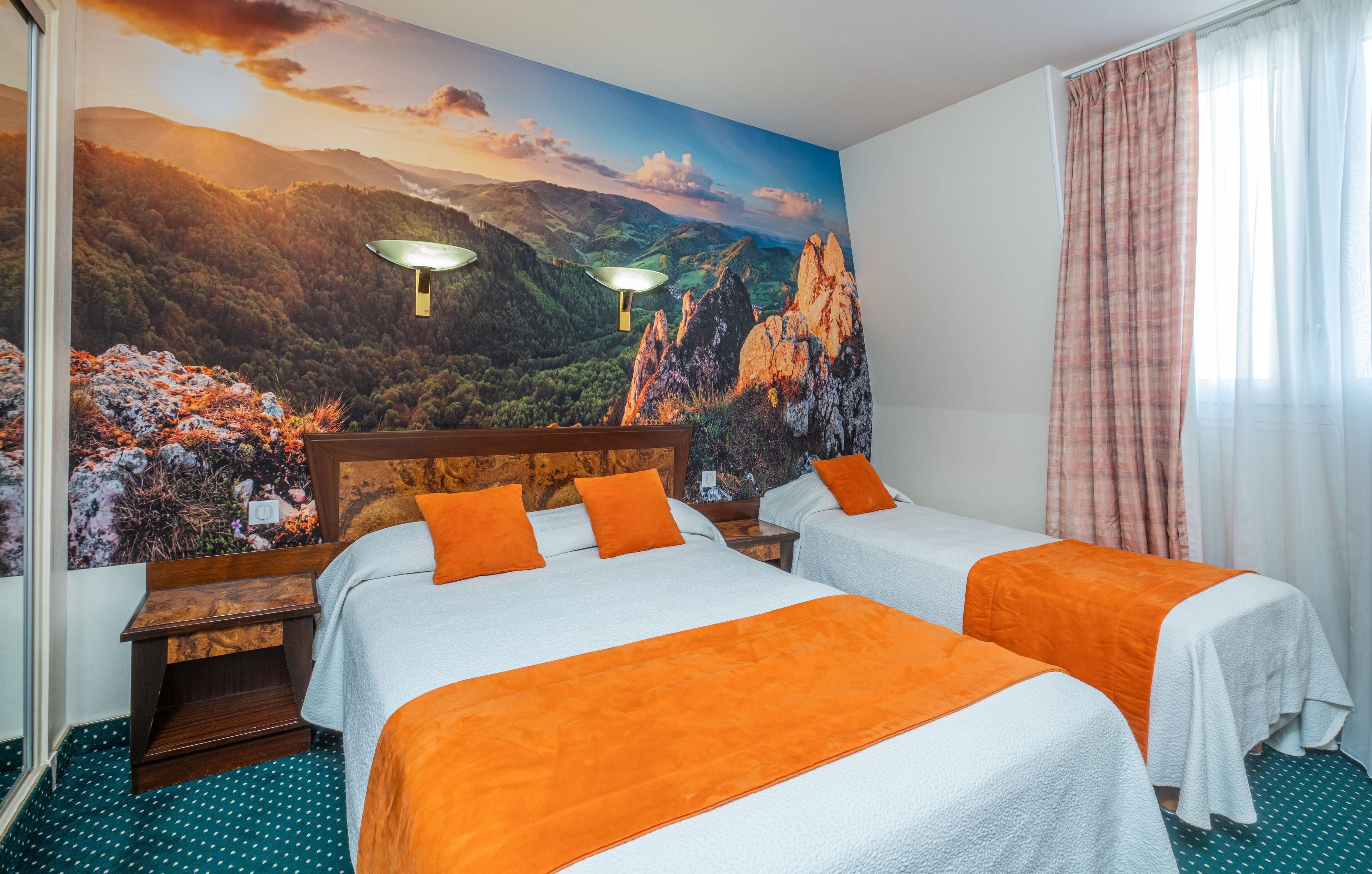 A hotel room with two beds, a large mural of a mountainous landscape, a wooden nightstand, and a window with curtains.