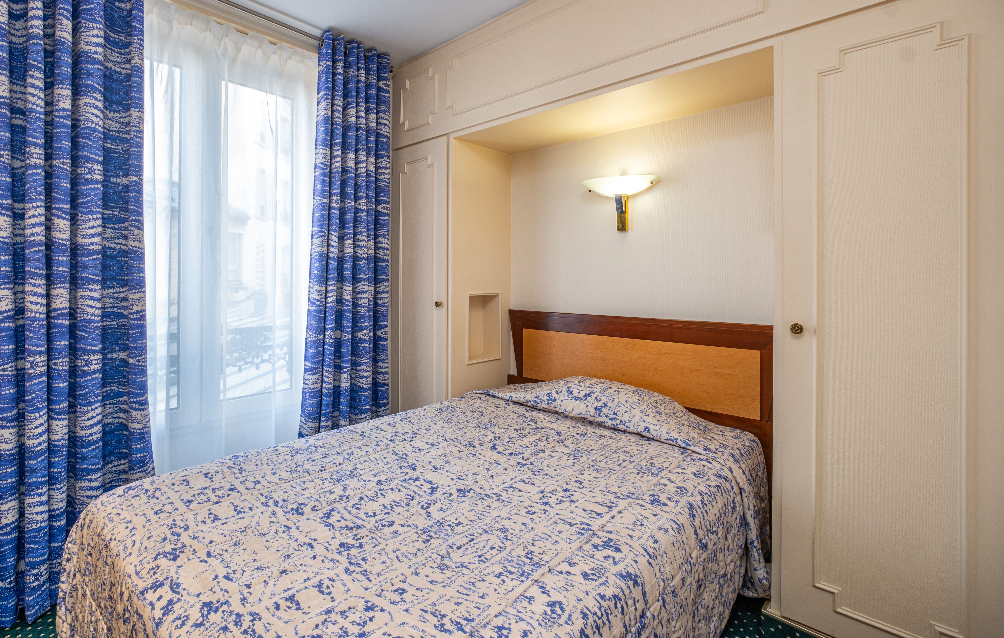 A hotel room with a bed, a nightstand, a wall-mounted lamp, and a window with curtains.
