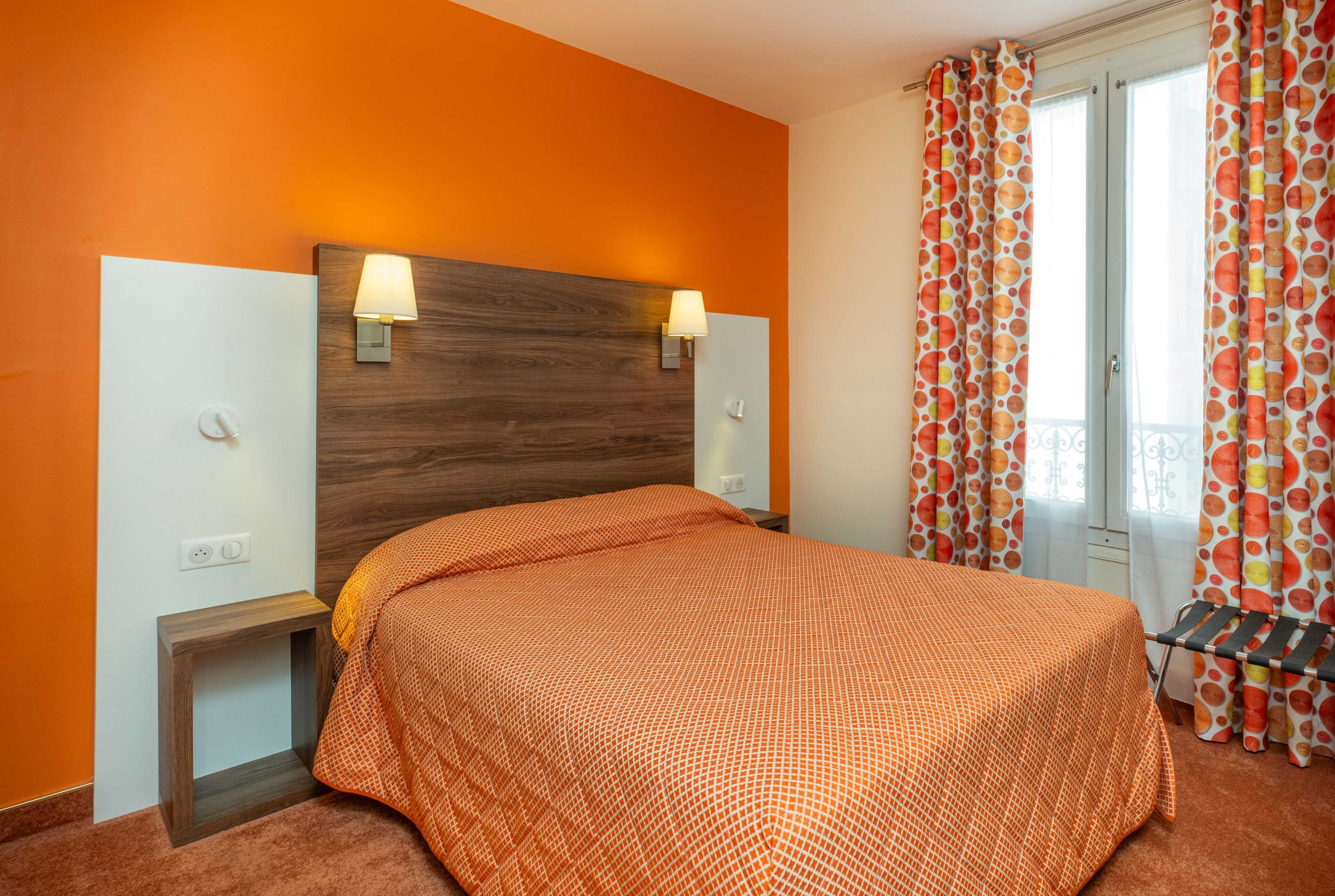 Quarto de hotel com cabeceira de madeira, duas luminárias de apoio, colcha de cama laranja com padrão e uma janela com cortinas coloridas.