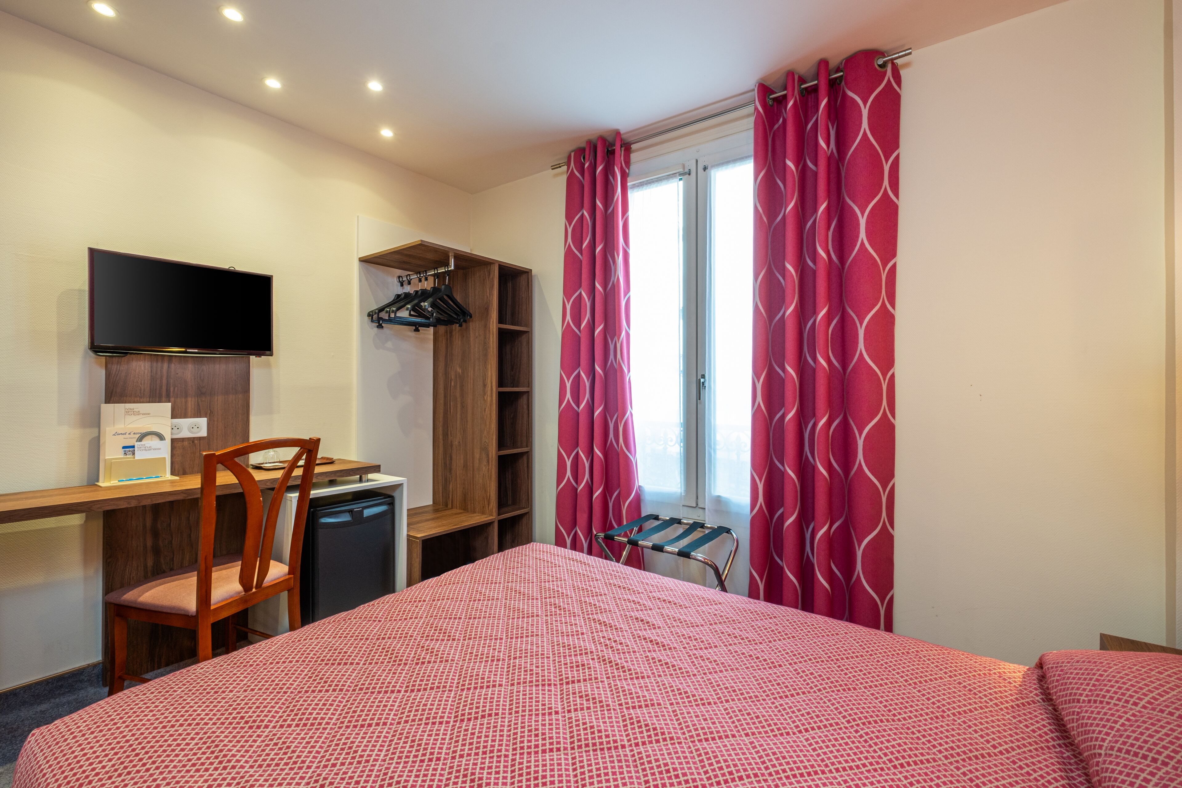 A hotel room with a bed, a desk with a chair, a television, a wardrobe, and a window with curtains.