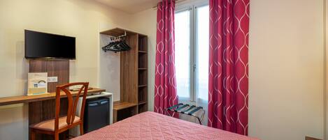 A hotel room with a bed, a desk with a chair, a television, a wardrobe, and a window with curtains.