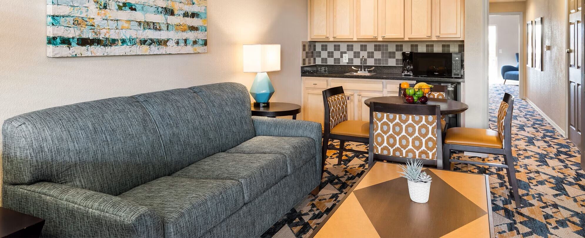 Sala de estar con sofá gris, una mesita con una planta y una zona de cocina con microondas y frutero.