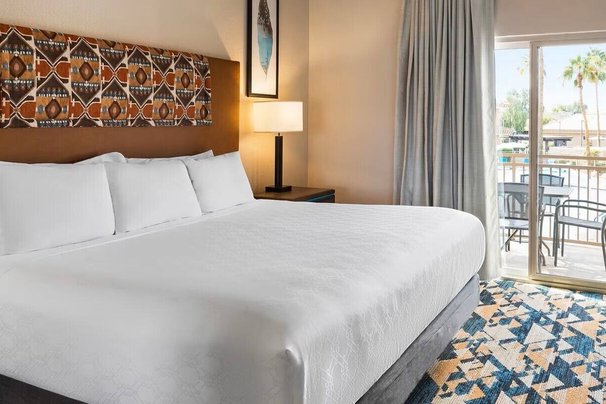 A hotel room with a large bed, a blue armchair, a patterned rug, and a view of the outdoor area through a sliding door.