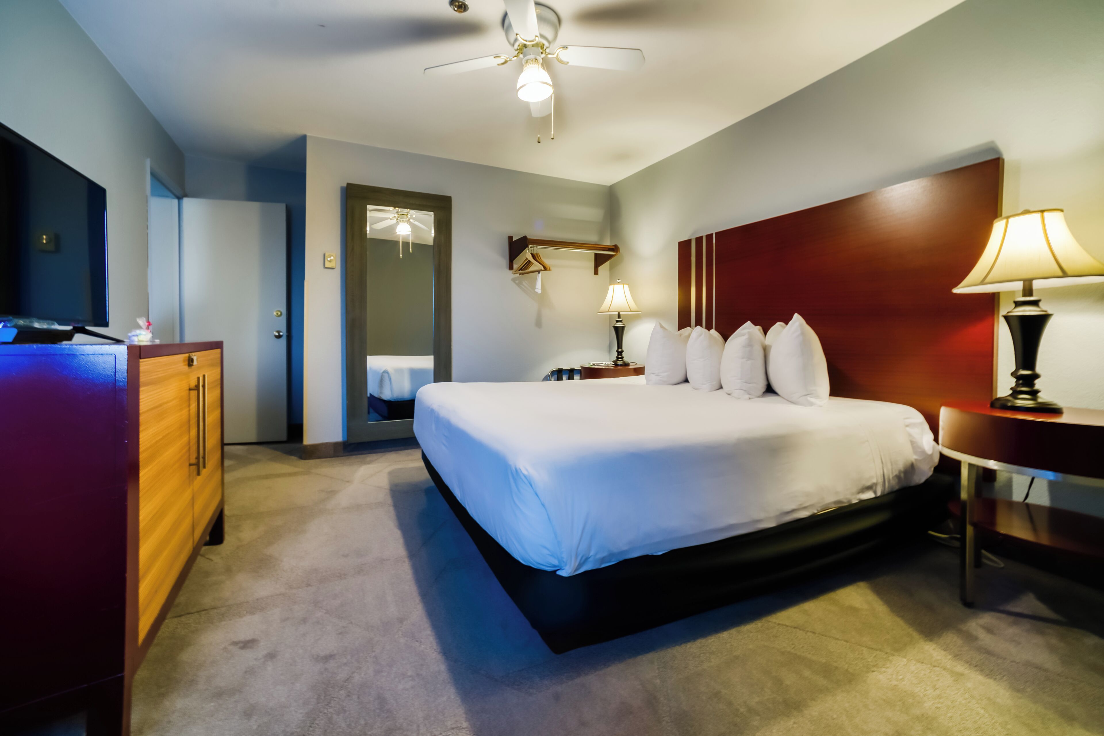 A hotel room with a large bed, a wooden dresser, a desk with a lamp, a ceiling fan, and a mirror.