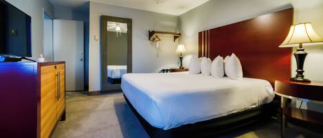 A hotel room with a large bed, a wooden dresser, a desk with a lamp, a ceiling fan, and a mirror.