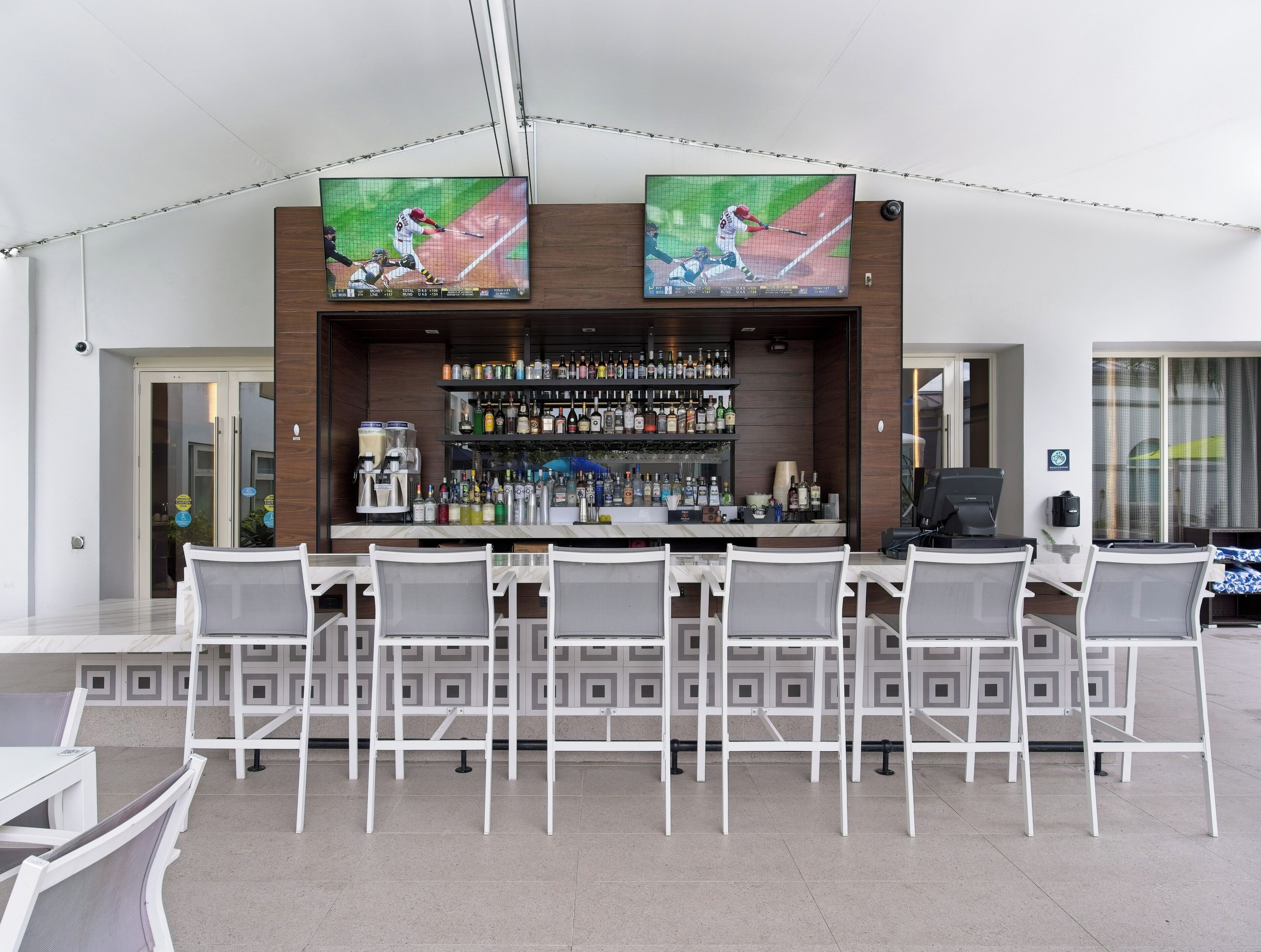 A bar area with a long counter, multiple chairs, and a wall-mounted TV screen displaying a baseball game.