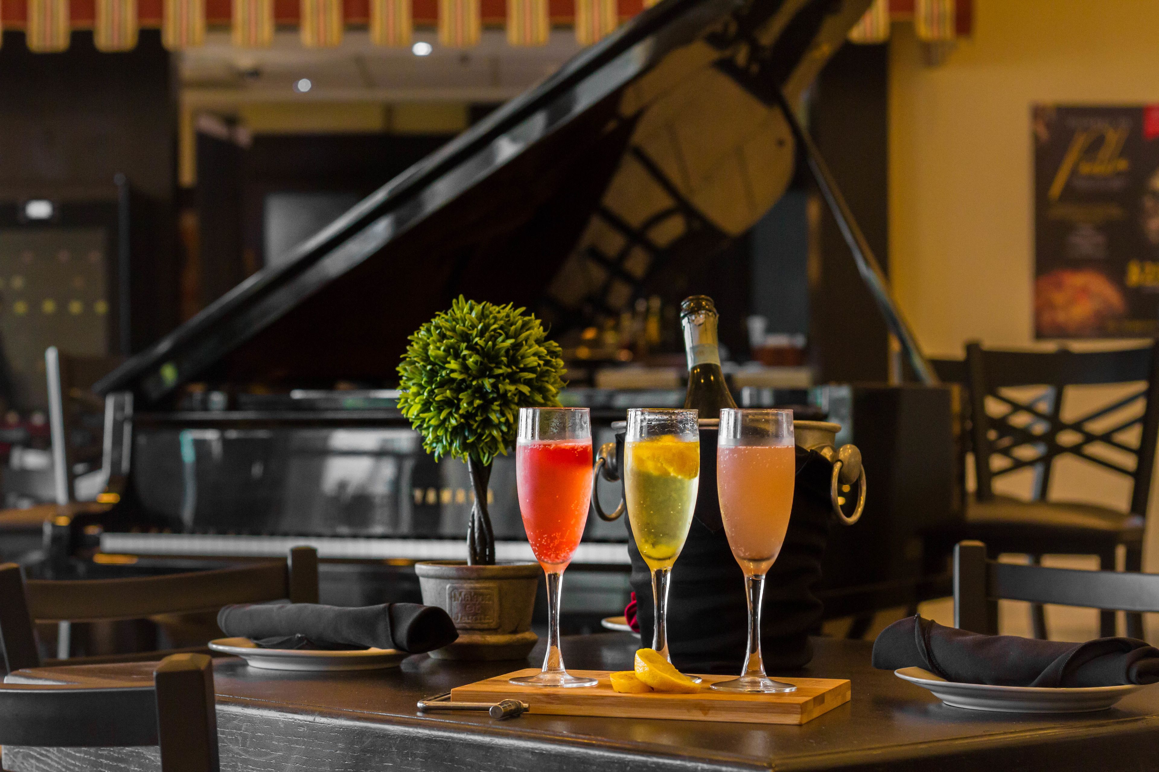 A bar area with a piano in the background, three cocktails on a wooden board, and a small potted plant.