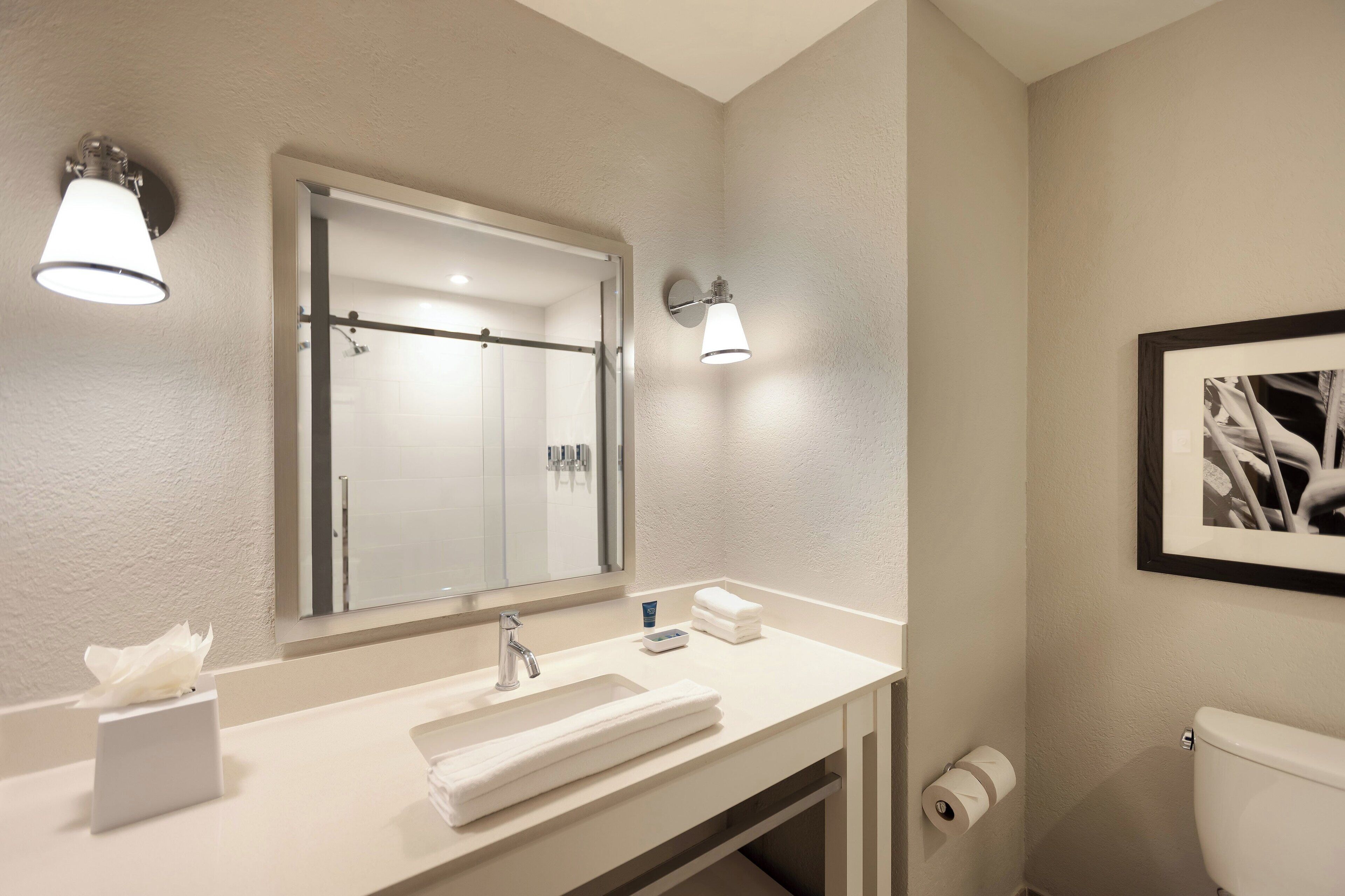 A bathroom with a sink, mirror, towel, toilet, and framed picture on the wall.